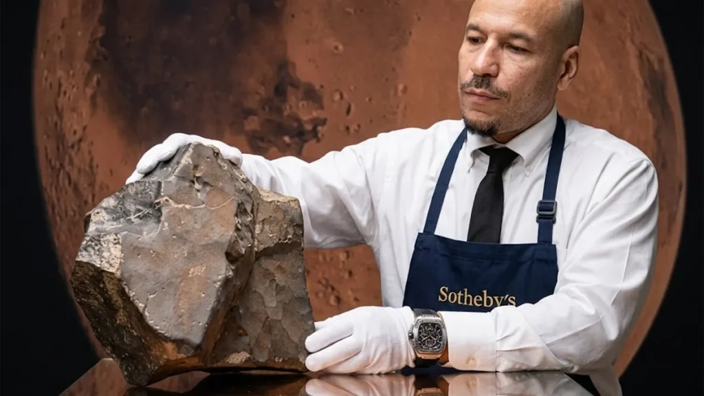 A high-resolution photograph of a Sotheby's specialist wearing white gloves and a navy apron, carefully handling the large, dark-toned NWA 16788 Martian meteorite. A large circular image of the planet Mars is visible in the background