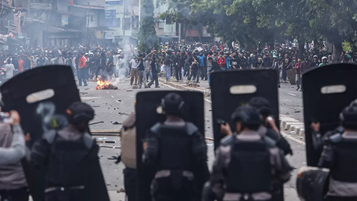 Riot police in black tactical gear and shields face off against protesters on a smoke-filled street in Jakarta during the August 2025 demonstrations.