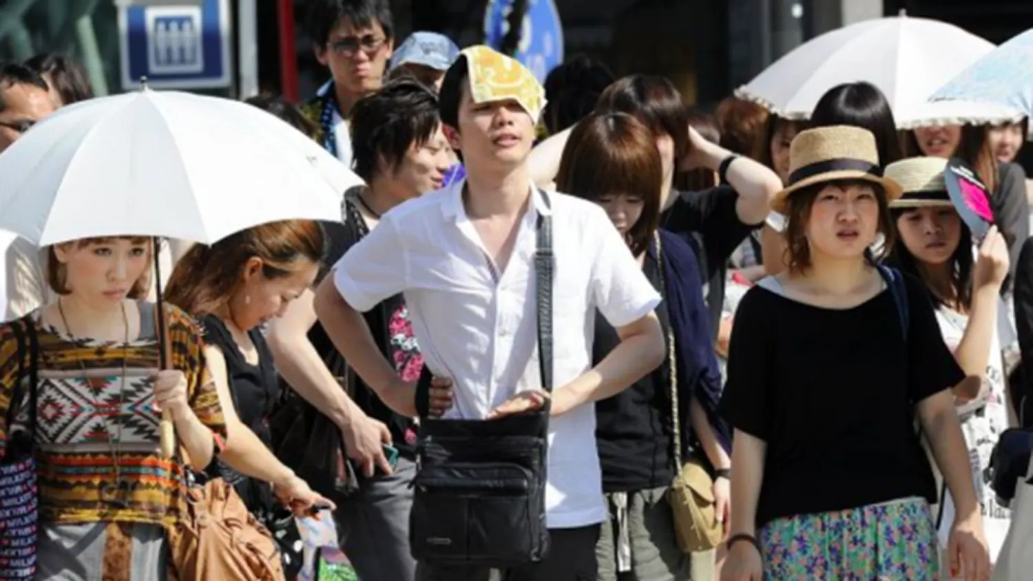 A dense crowd of people in East Asia walking outside on a extremely hot day. A central figure is wiping sweat from his forehead with a yellow cooling towel. Others are seen using white umbrellas, straw hats, and hand fans for shade