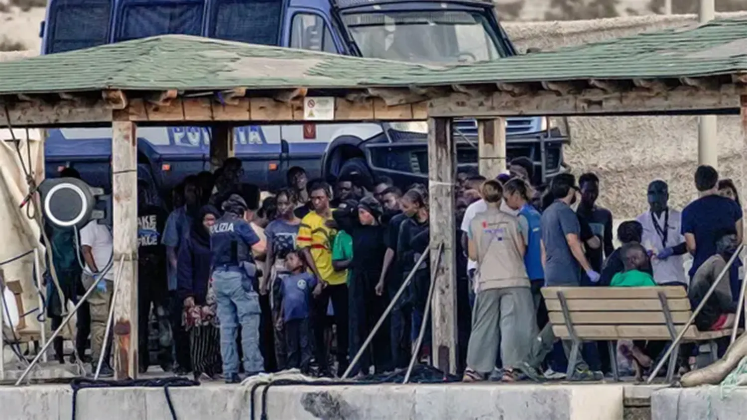 A group of migrants and refugees standing under a wooden shelter at a Lampedusa port being assisted by Italian police and humanitarian aid workers following a boat capsizing in 2025