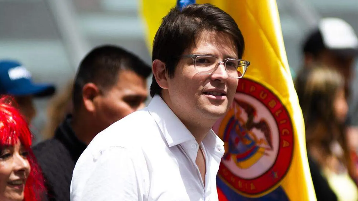Close-up of Colombian Senator and 2026 presidential pre-candidate Miguel Uribe Turbay wearing a white shirt and glasses, smiling in front of a Colombian flag during a 2025 campaign event