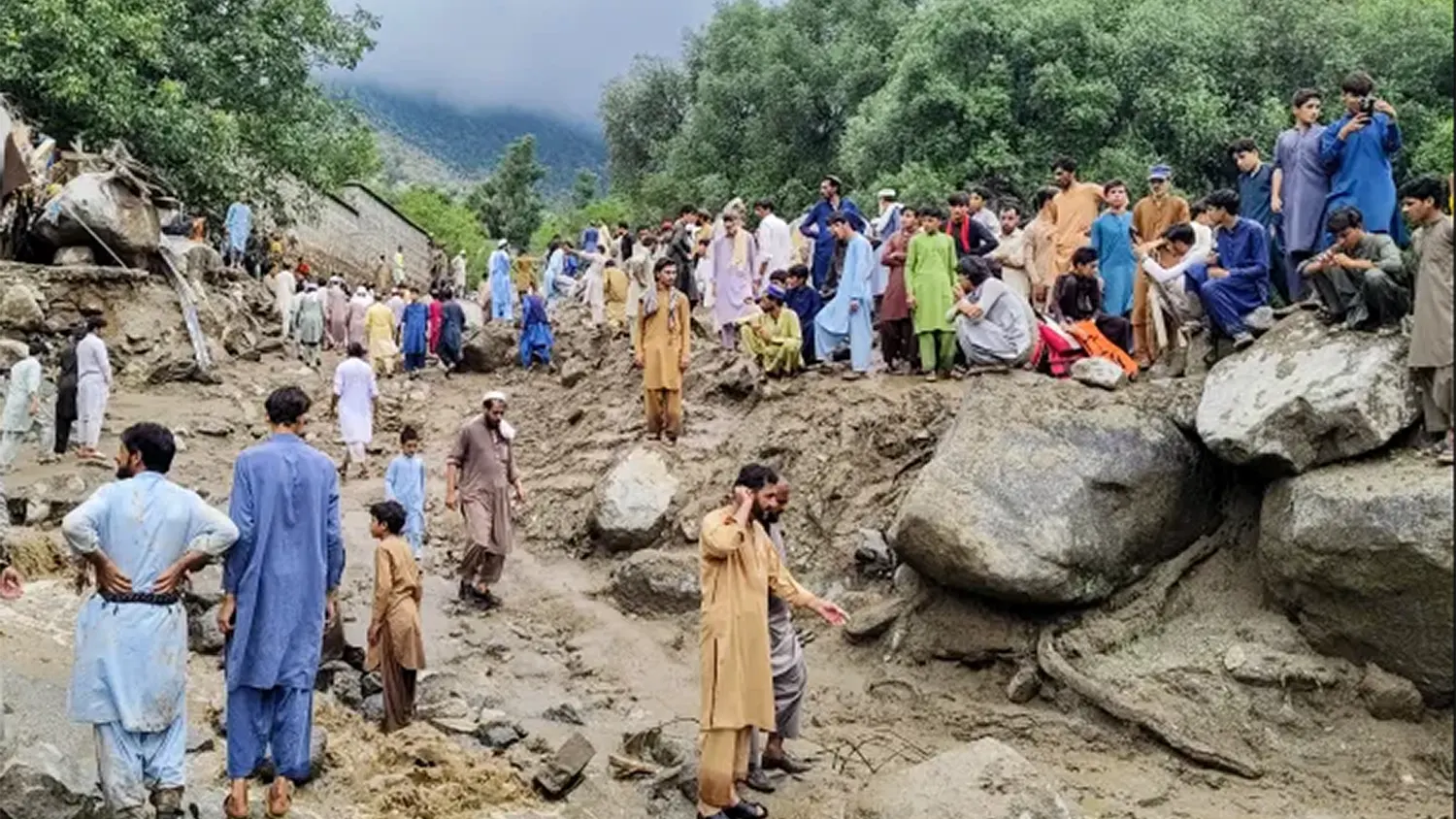 Displaced residents standing among large boulders and mud debris in a flood-ravaged valley in Pakistan during the catastrophic August 2025 monsoon season.