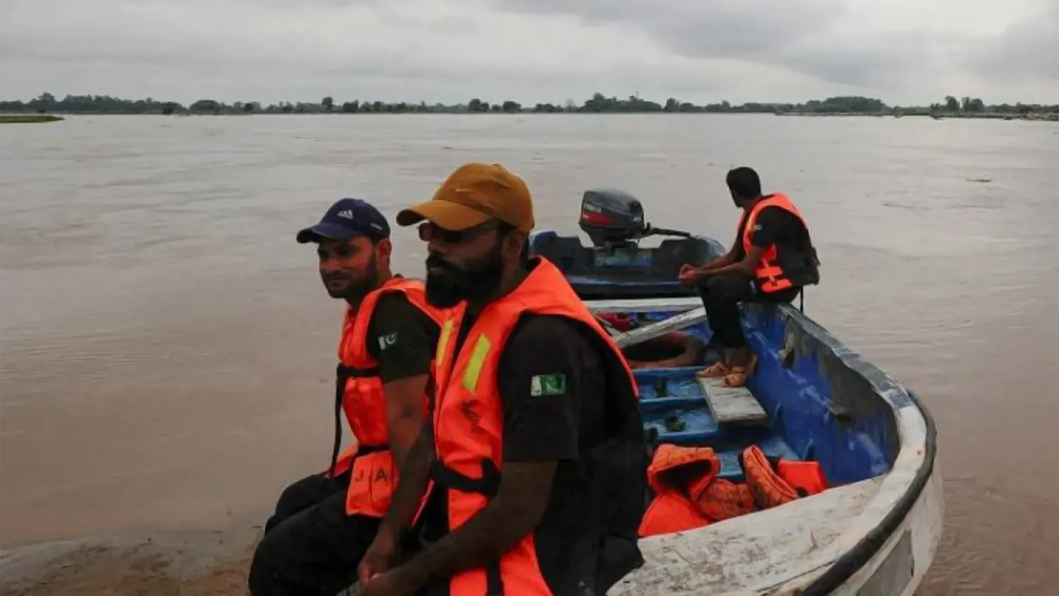 Three rescue workers in orange life vests sit on a blue motorboat on the flooded Sutlej River in Eastern Pakistan during mass evacuations.