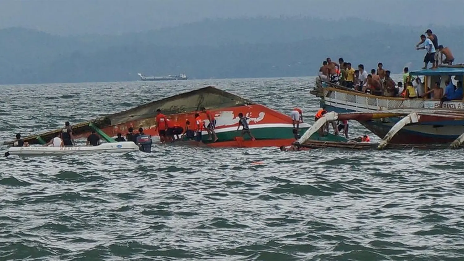 A crowded rescue scene on the water with two motorized outrigger boats, one partially capsized, as many people scramble for safety while a rescue team member in an orange vest approaches in a small white boat. In the background, there is a hazy, mountainous landscape.