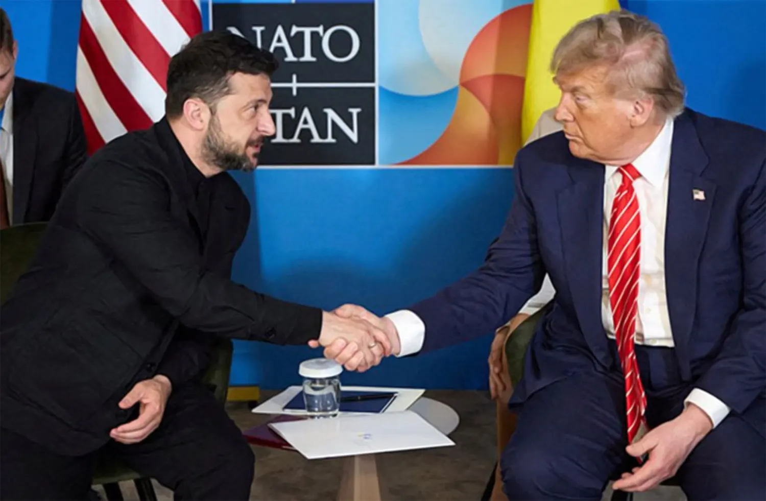 President Volodymyr Zelenskyy and President Donald Trump shaking hands during a diplomatic meeting in Washington D.C. with NATO and international flags in the background.