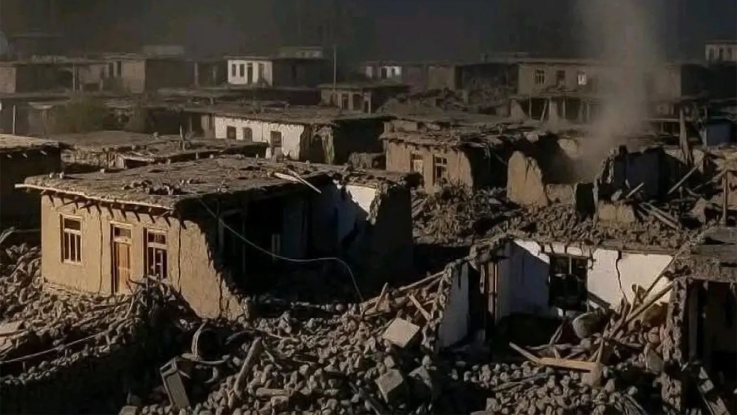 A landscape view of a village in eastern Afghanistan showing extensive destruction from an earthquake. Mud-brick houses are partially collapsed, with rubble, wooden beams, and dust filling the narrow streets. A light haze or smoke hangs in the air over the damaged settlement