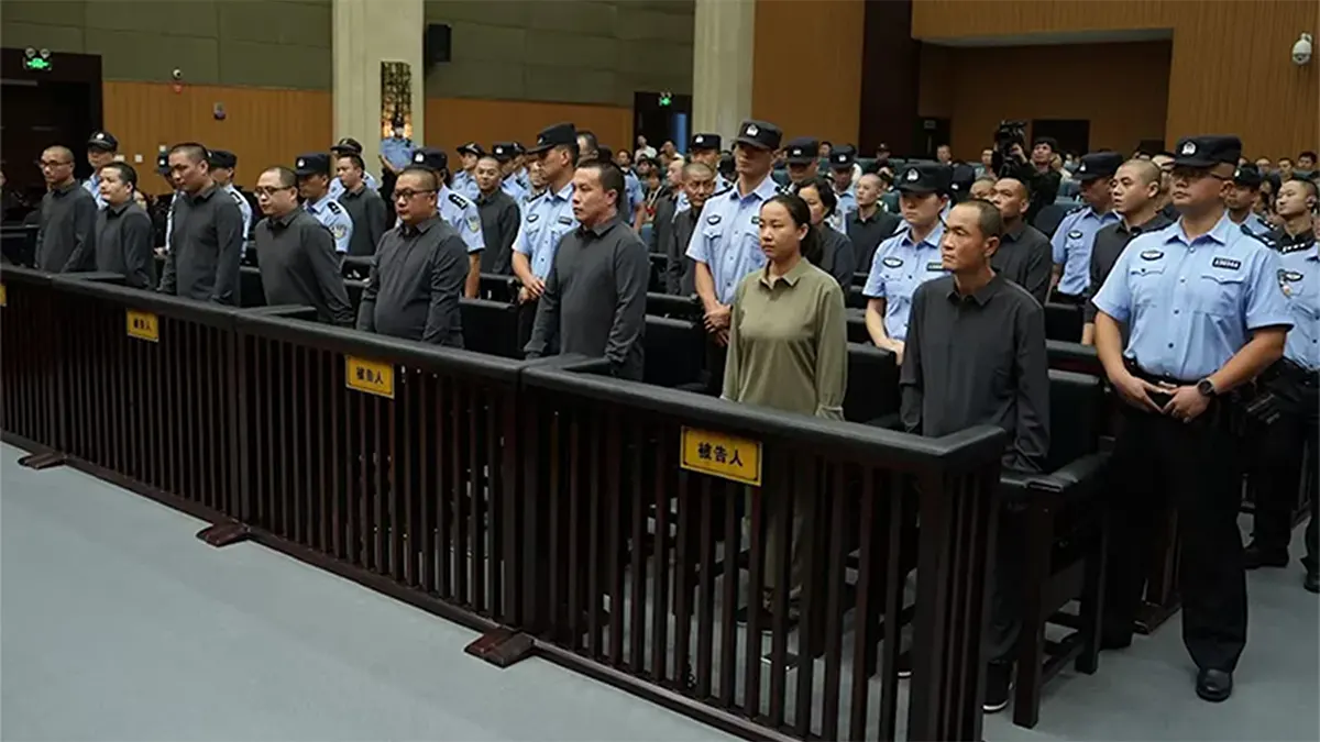 A crowded courtroom in China where a line of defendants in plain clothes stands behind a dark wooden barrier labeled with yellow signs. Numerous police officers in light blue uniforms stand guard behind and beside the defendants during a sentencing hearing.
