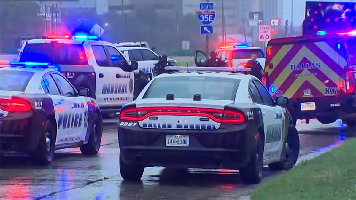 Multiple emergency vehicles, including a Dallas Marshal squad car, a Dallas Police vehicle, and a Dallas Fire-Rescue truck, are parked on a wet road with their red and blue emergency lights flashing near an interstate highway sign.