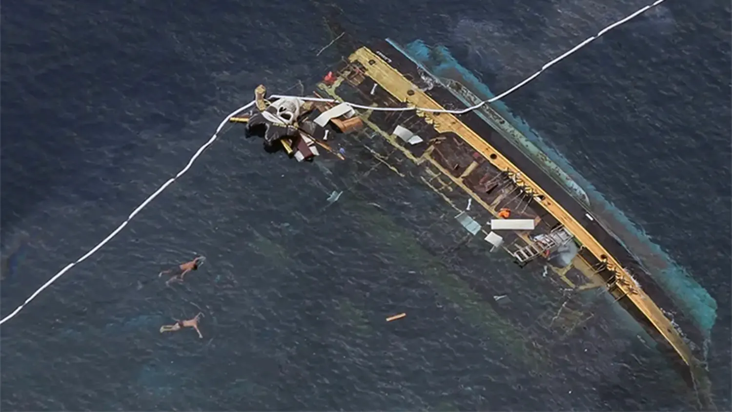Aerial view of a large wooden whaleboat capsized and partially submerged in the Congo River near Malange, DRC, with floating debris and oil sheen visible on the water surface.
