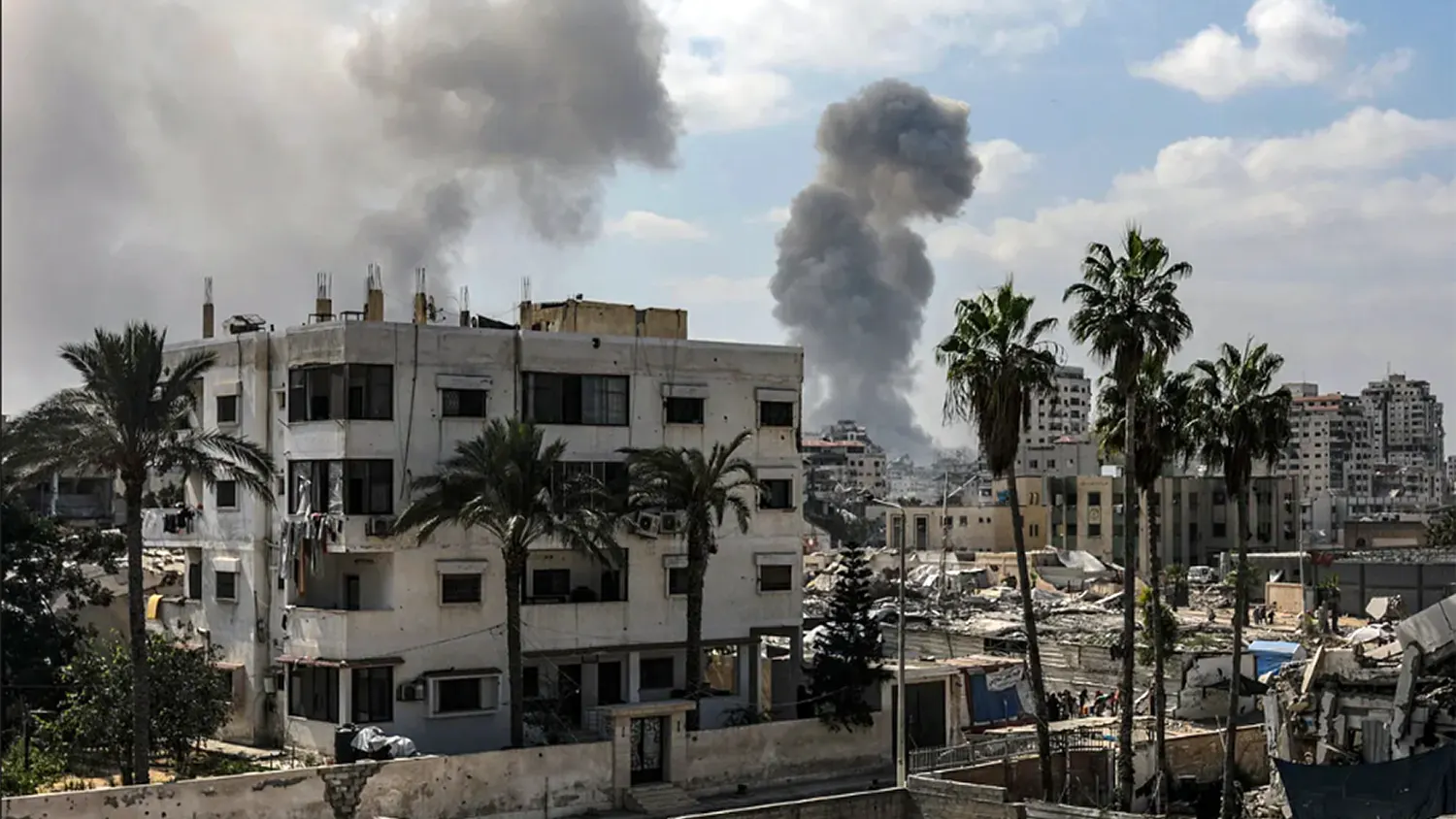 Large plumes of dark grey smoke rising above residential buildings and palm trees in Gaza City during the Israeli ground offensive on September 17, 2025