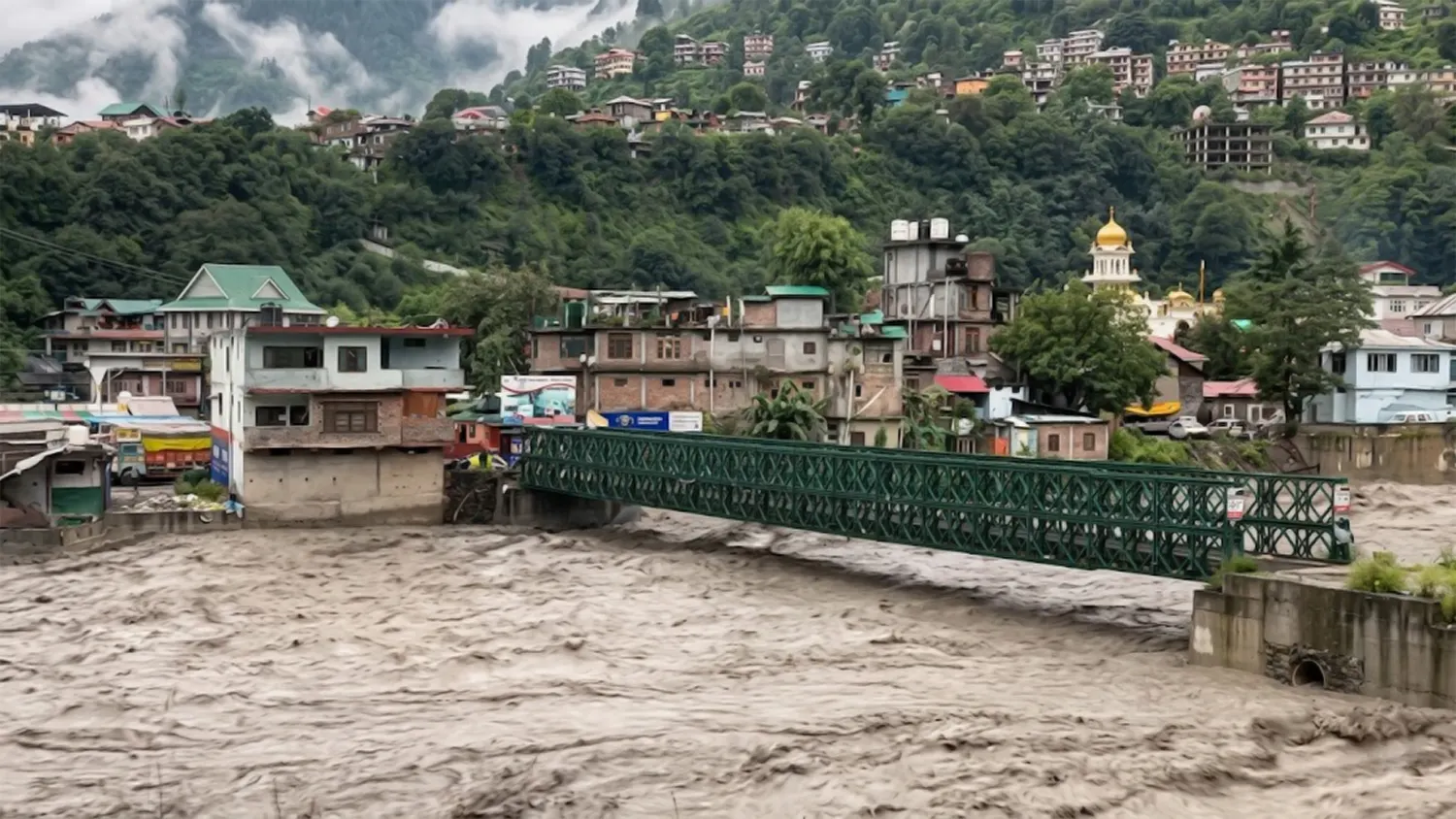 A high-angle view of Kullu, Himachal Pradesh, showing a bridge over the extremamente turbulent Beas River during a severe monsoon flood. Multistory buildings line the banks with a temple visible in the background