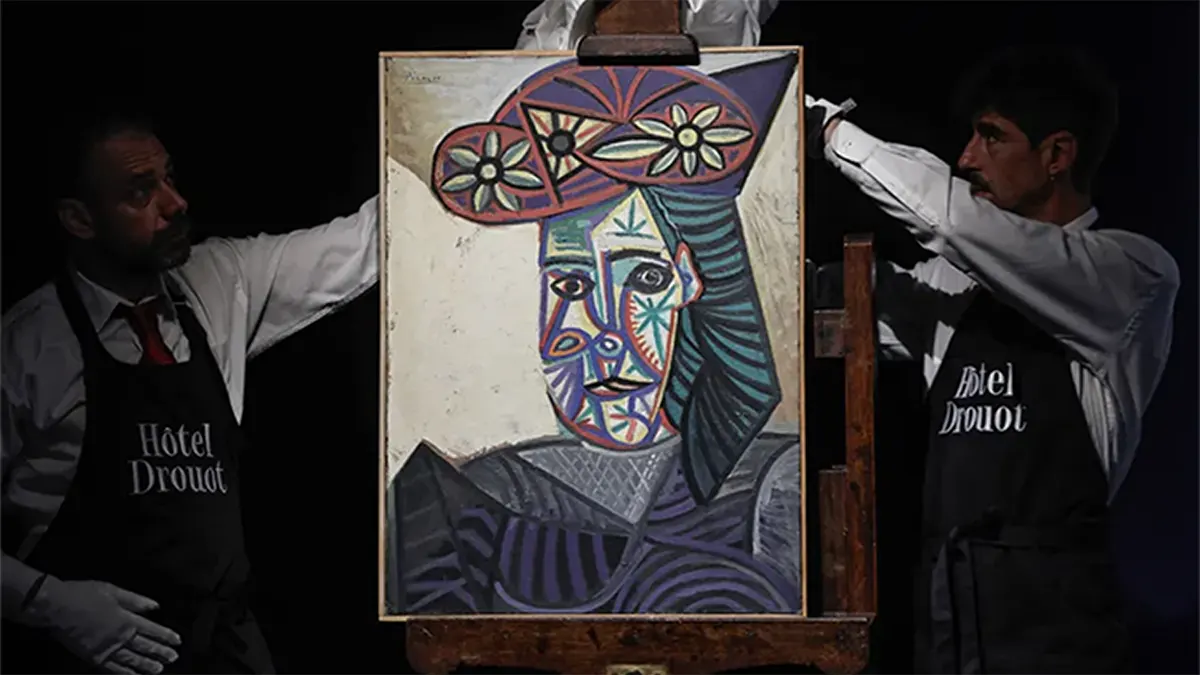 Two handlers in white shirts and black Hôtel Drouot aprons carefully holding and displaying the rediscovered 1943 Cubist oil painting 'Bust of a Woman with a Flowery Hat' by Pablo Picasso on a wooden easel.