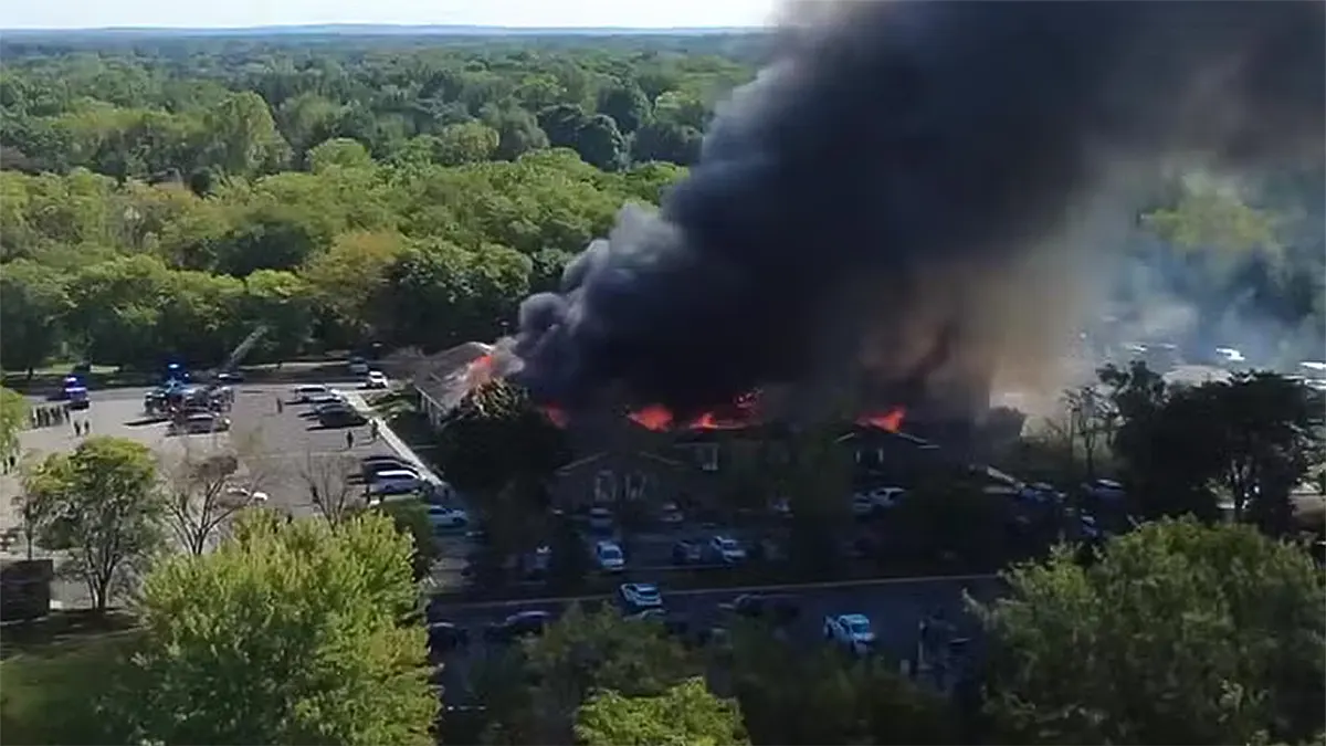An aerial view of a church building engulfed in large orange flames with a massive plume of thick black smoke rising into the sky, surrounded by emergency vehicles and parked cars in Michigan.