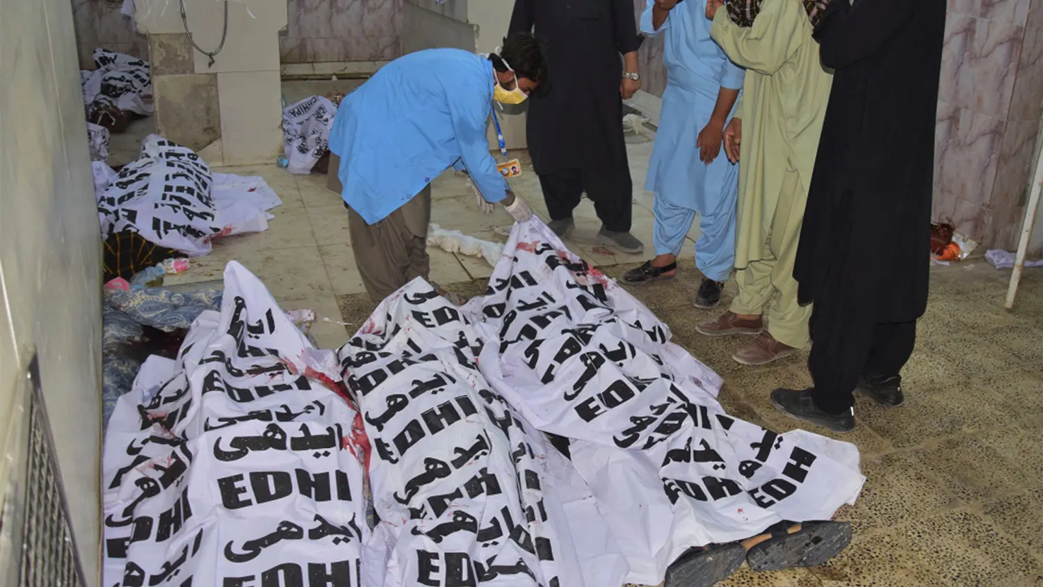 Mourning men stand over several bodies covered in white Edhi foundation shrouds in a morgue or hospital setting following a suicide bombing in Quetta, Balochistan.