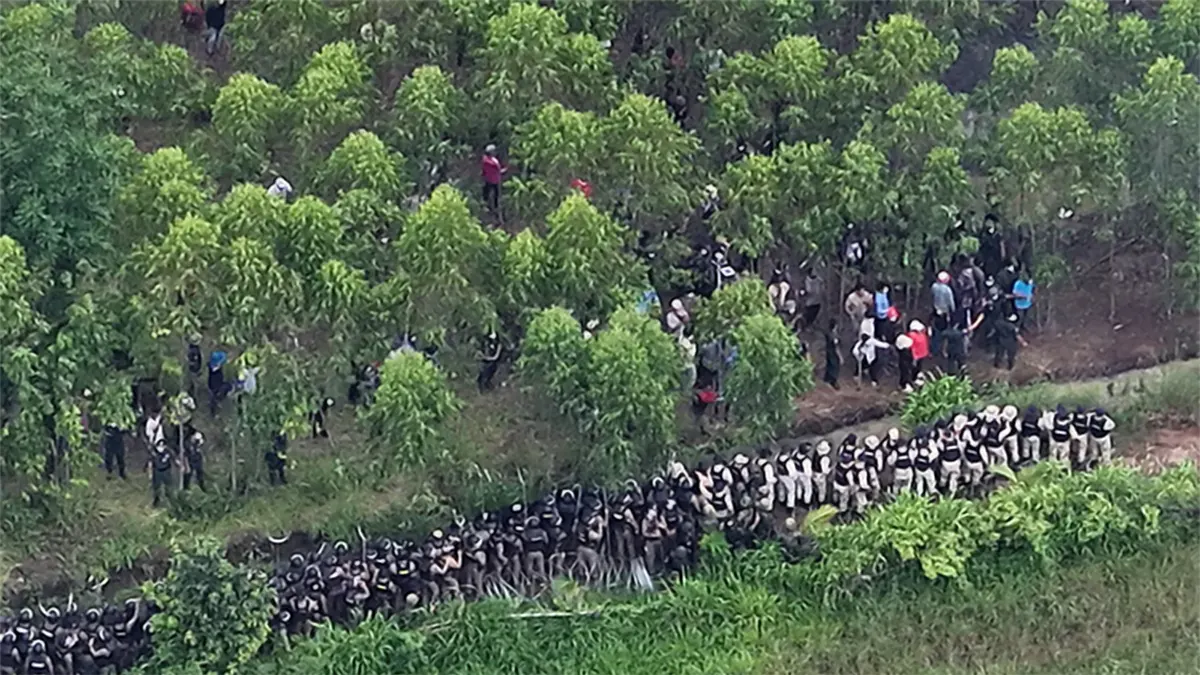 Aerial view of a long line of anti-riot police in dark uniforms and helmets standing behind a barrier, confronting a large crowd of civilian protesters scattered among a dense grove of green trees at the disputed Thai-Cambodian border village area