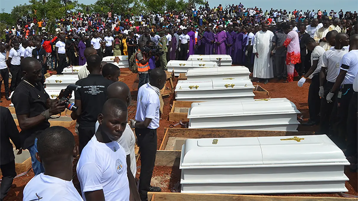 A mass funeral service in Nigeria featuring rows of white coffins being prepared for burial as Christian clergy in white and purple robes stand before a large crowd of mourners.