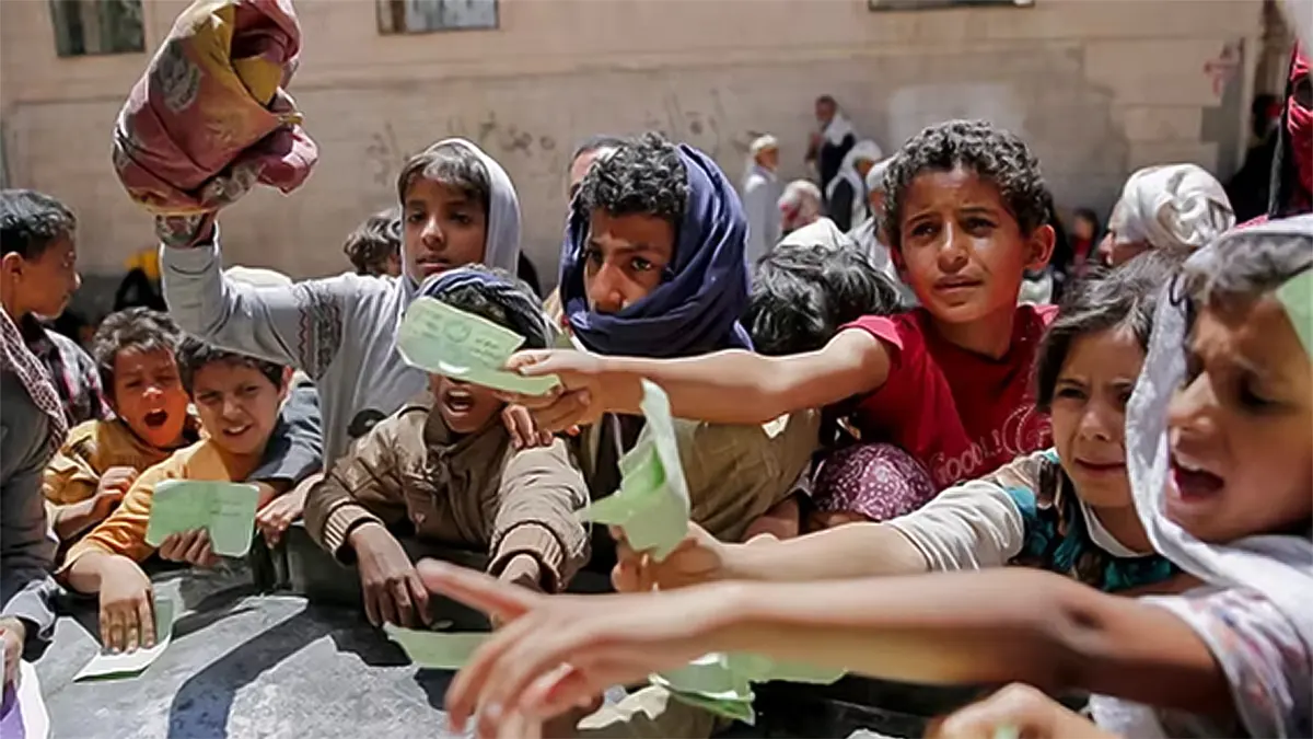 A group of young Yemeni children behind a concrete barrier with their hands reaching out for official aid vouchers or paper notes during a food distribution event in San’a, Yemen, in 2025.