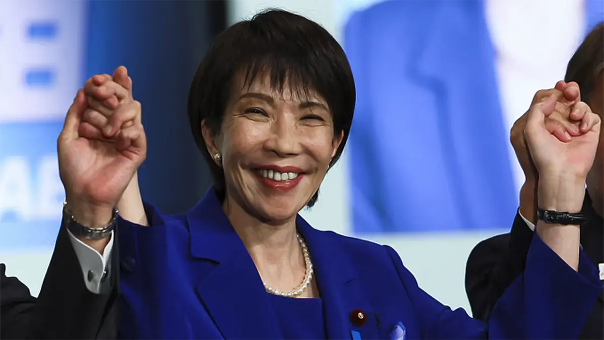 Close-up portrait of a smiling Sanae Takaichi in a bright blue blazer and pearl necklace, holding hands aloft in victory with party members following her election win.