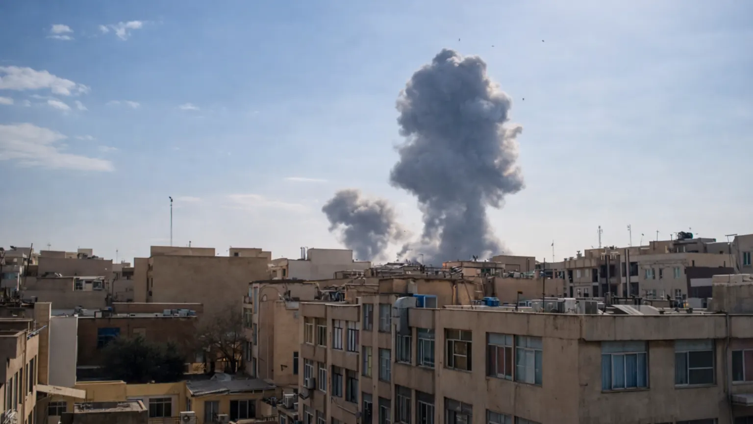 Large smoke plumes rising over Tehran skyline after reported airstrikes, viewed from residential buildings under a clear blue sky