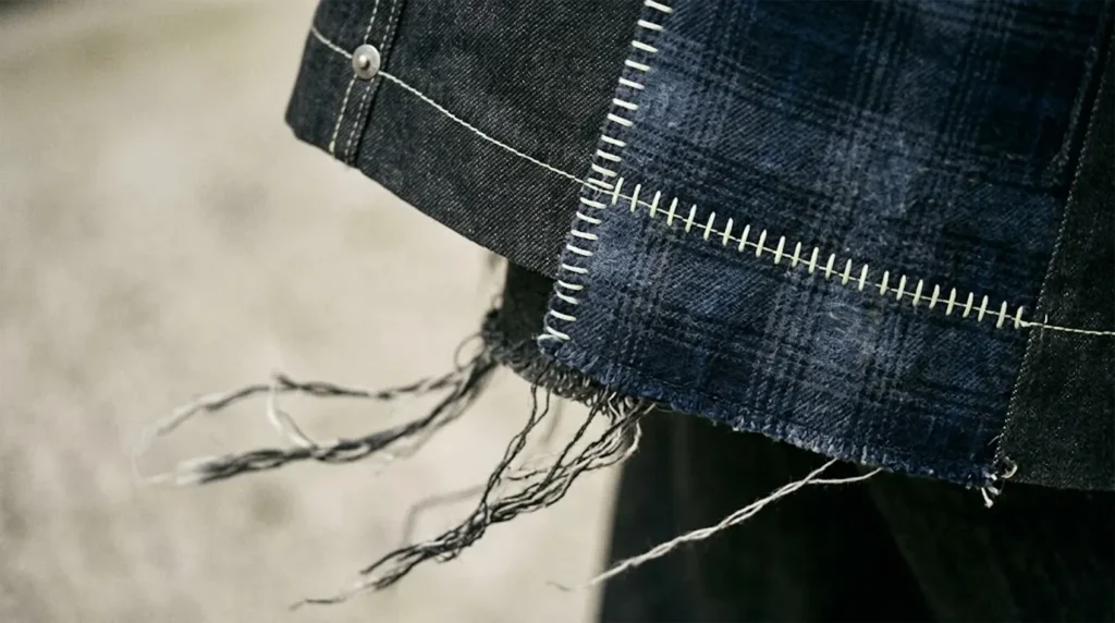 Extreme close-up of a deconstructed dark denim jacket featuring off-white neon contrast stitching, a blue plaid patchwork repair, and a frayed raw hem with loose threads.