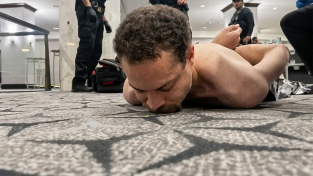 A high-resolution photo of suspect Cole Tomas Allen being apprehended by law enforcement on the carpeted floor of the Washington Hilton. He is shirtless, handcuffed behind his back, and held down by officers following a security breach at the 2026 White House Correspondents' Dinner.