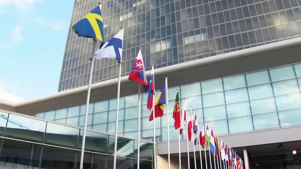 A row of European Union member state flags, including Sweden, Finland, and Slovakia, flying in front of the modern glass facade of the European Commission headquarters in Brussels.