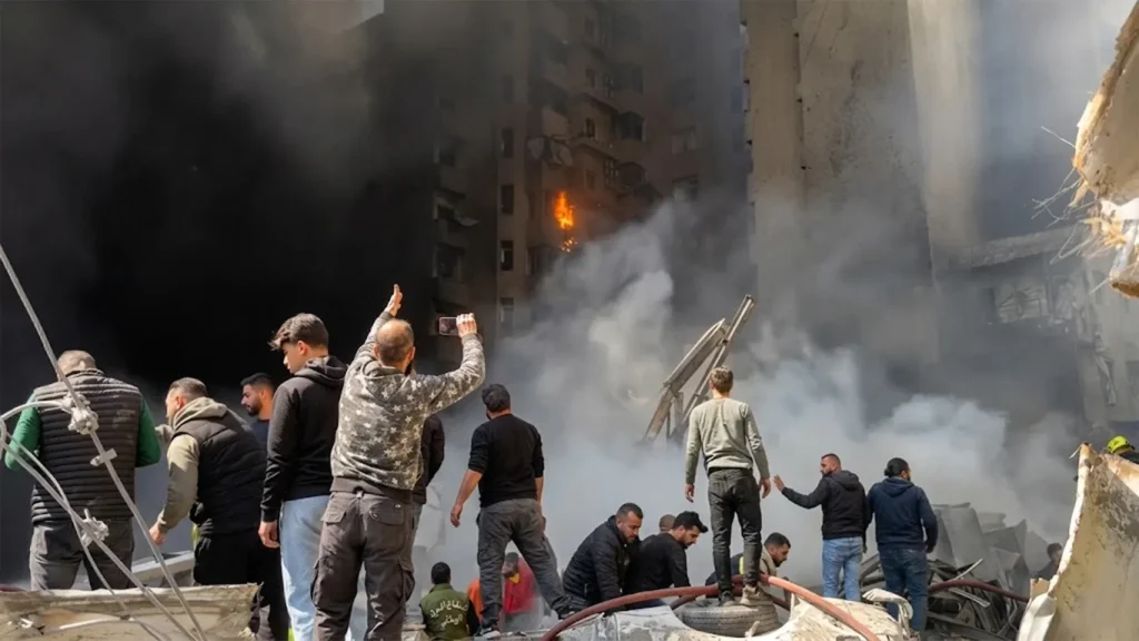 A high-resolution image showing a residential building on fire in the al-Mazraa district of Beirut. Thick smoke billows from the structure as civilians and rescue workers stand amidst rubble and debris in the foreground
