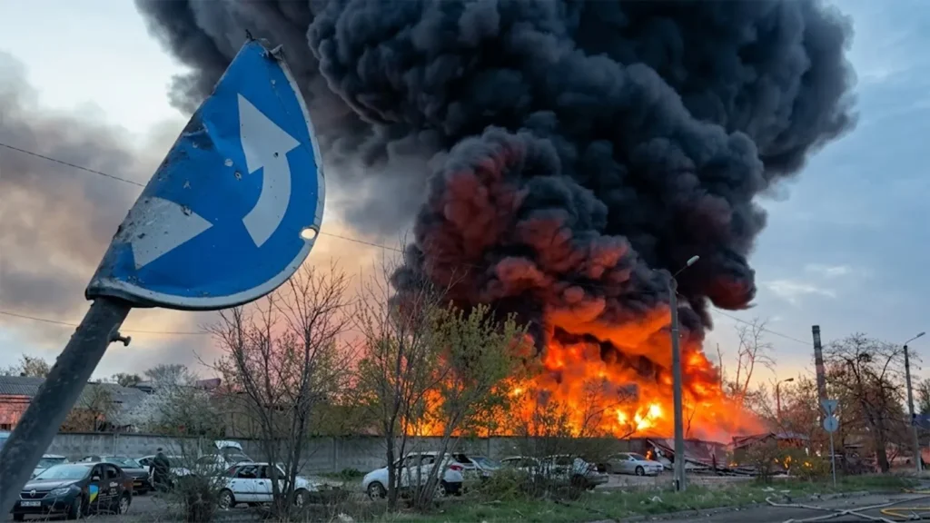 A massive plume of thick black smoke and intense orange flames rising from a hit building in Kyiv, Ukraine, following a large-scale Russian missile and drone assault on April 16, 2026. A damaged blue road sign is visible in the foreground