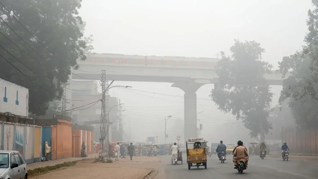 A high-resolution wide-angle view of a smog-filled street in Lahore, Pakistan, featuring an elevated Orange Line metro train bridge, rickshaws, and motorcycles obscured by thick grey haze