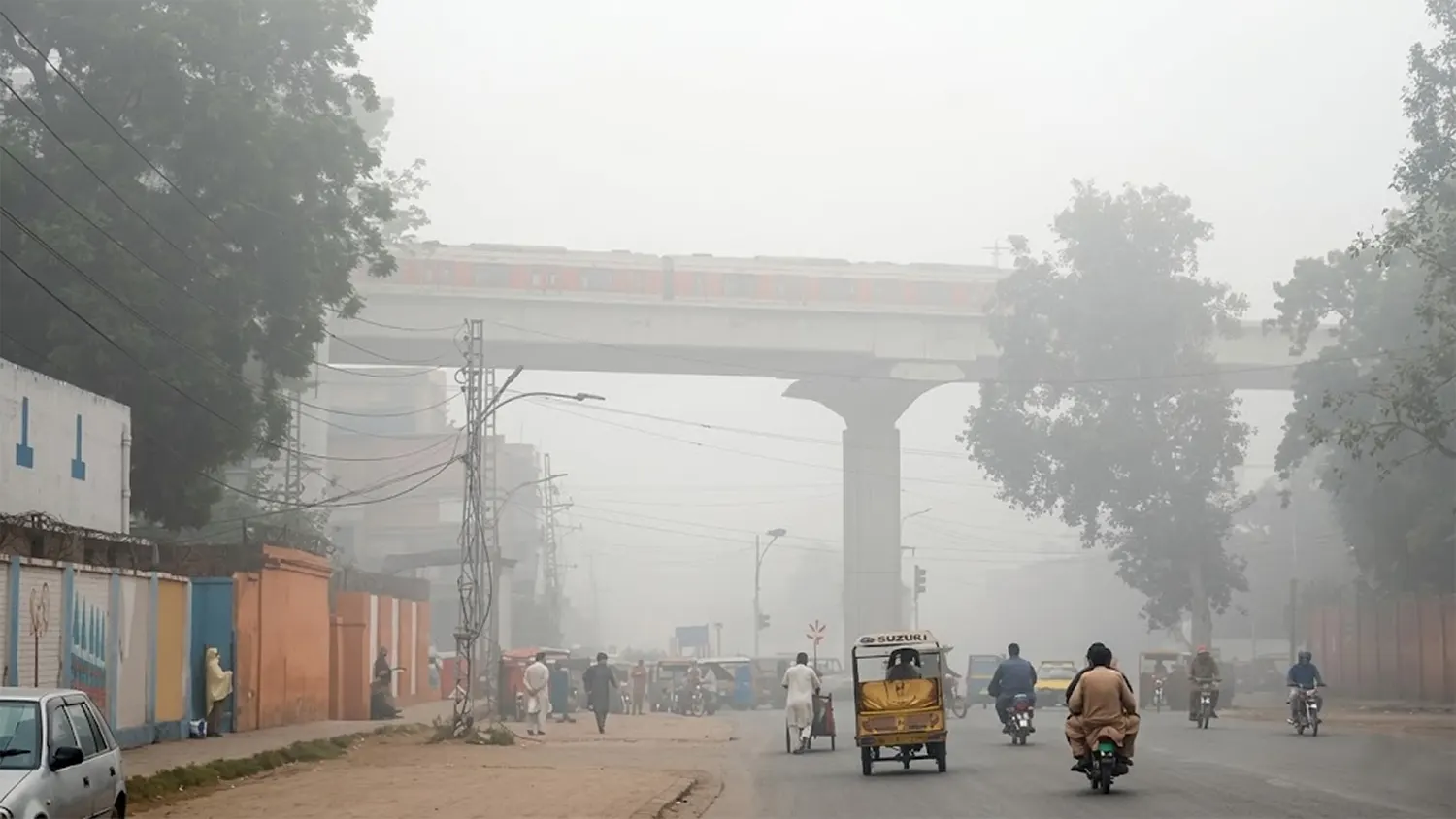 A high-resolution wide-angle view of a smog-filled street in Lahore, Pakistan, featuring an elevated Orange Line metro train bridge, rickshaws, and motorcycles obscured by thick grey haze