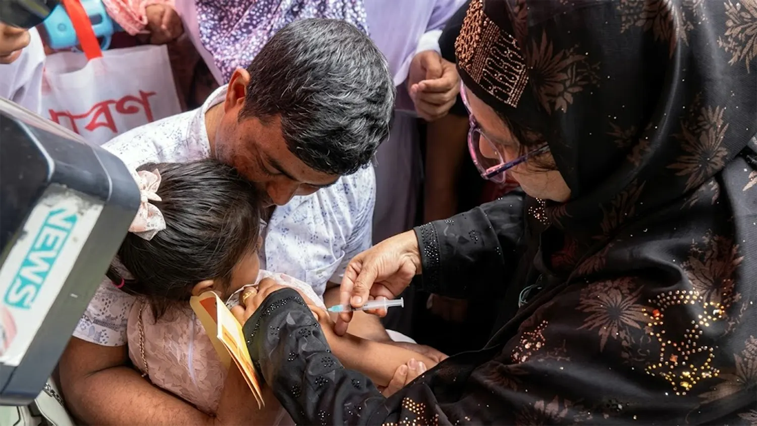 A health worker in a black hijab administers a measles vaccine to a young girl held by her father during a public health campaign in Bangladesh