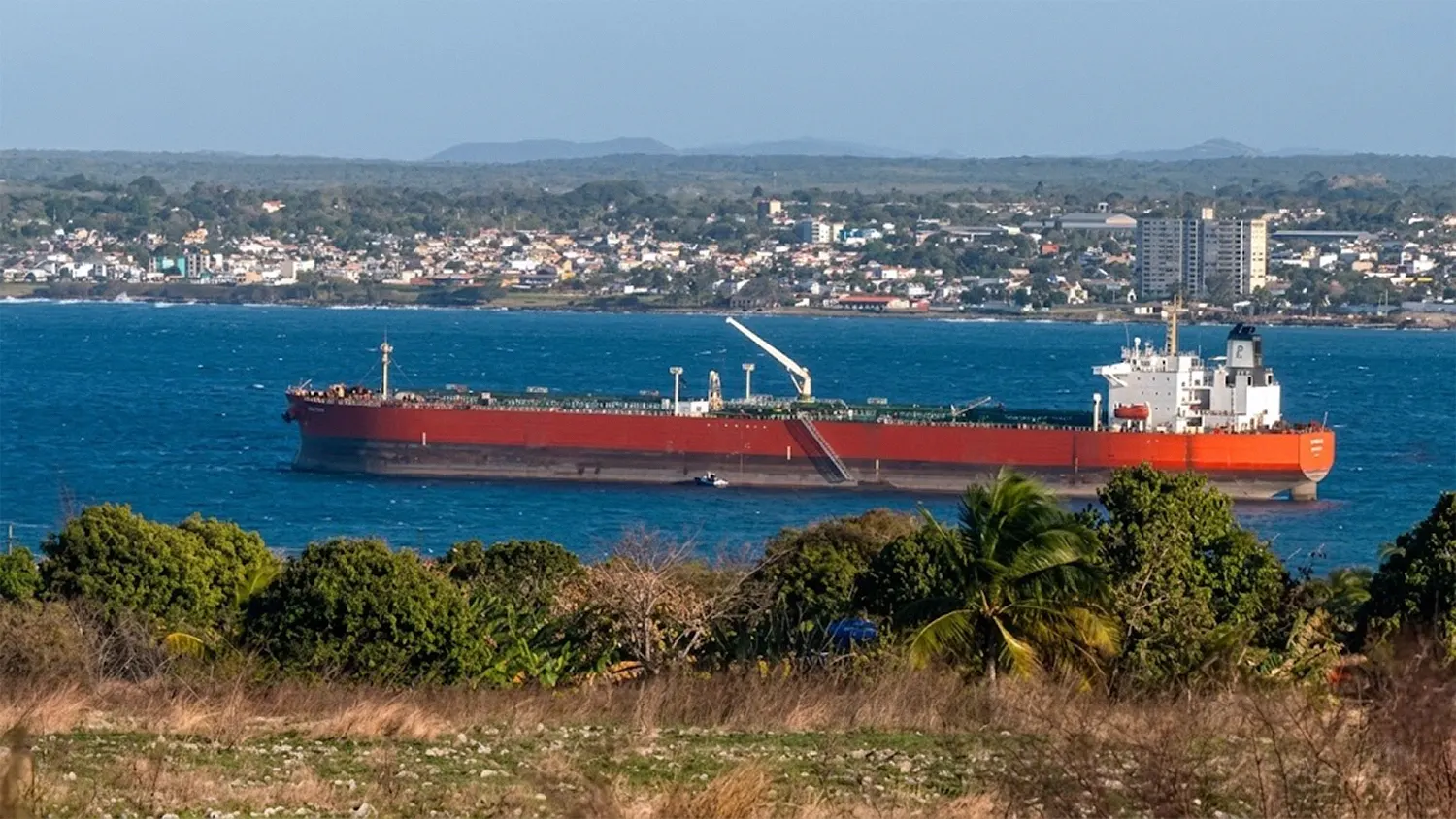 Large red and white Russian oil tanker, the Anatoly Kolodkin, anchored in the bay of Matanzas, Cuba, with the city skyline and tropical foliage in the foreground. High-resolution telephoto shot