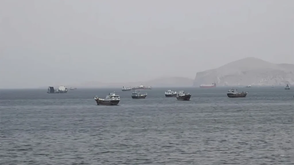 A wide-angle shot of multiple dhows and international cargo ships anchored or stalled in the hazy waters of the Strait of Hormuz during the 2026 maritime energy crisis.