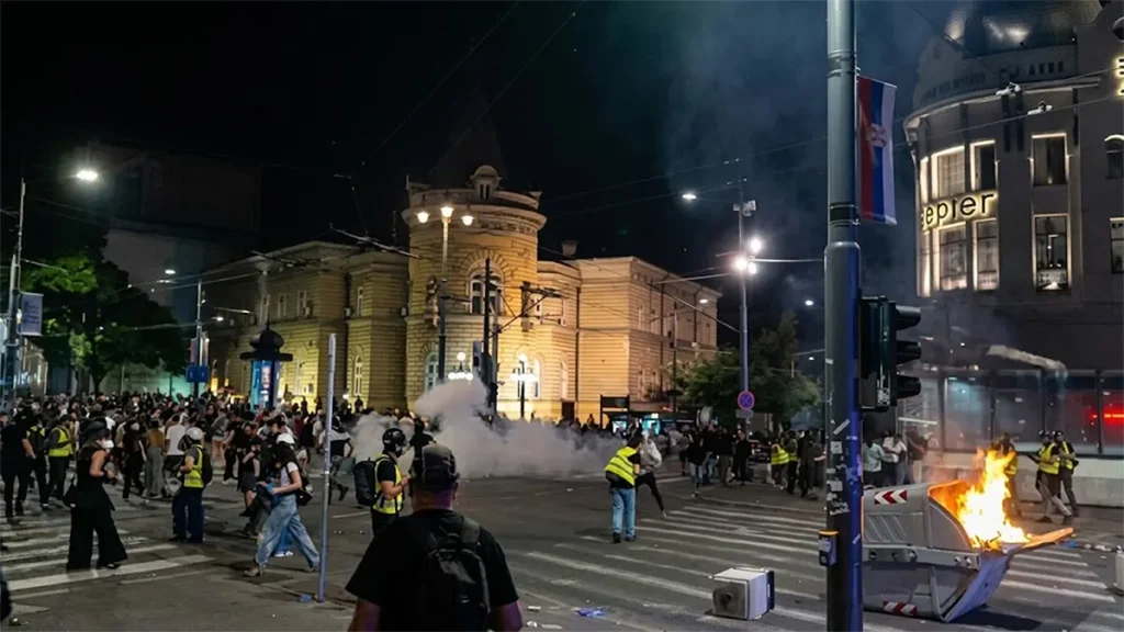 A night photograph of street clashes during a protest in Belgrade, Serbia, featuring a burning dumpster on a crosswalk, smoke or tear gas in the air, and demonstrators moving near historic buildings