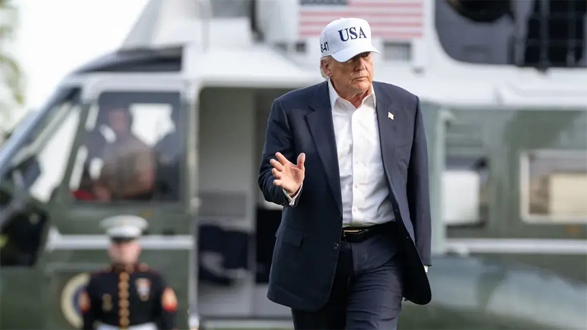 President Donald Trump wearing a navy blue suit and a white "USA" baseball cap while walking across the South Lawn with a Marine Corps helicopter (Marine One) in the background