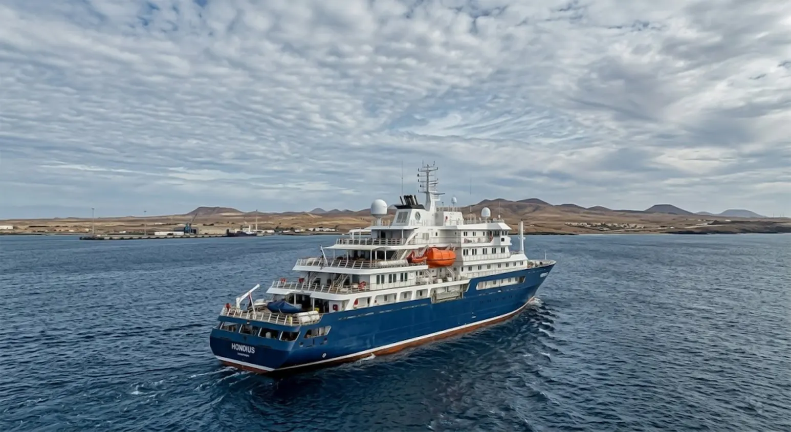 A high-angle professional photograph of the blue and white expedition cruise ship MV Hondius sailing on deep blue water near a desert coastline under a cloudy sky