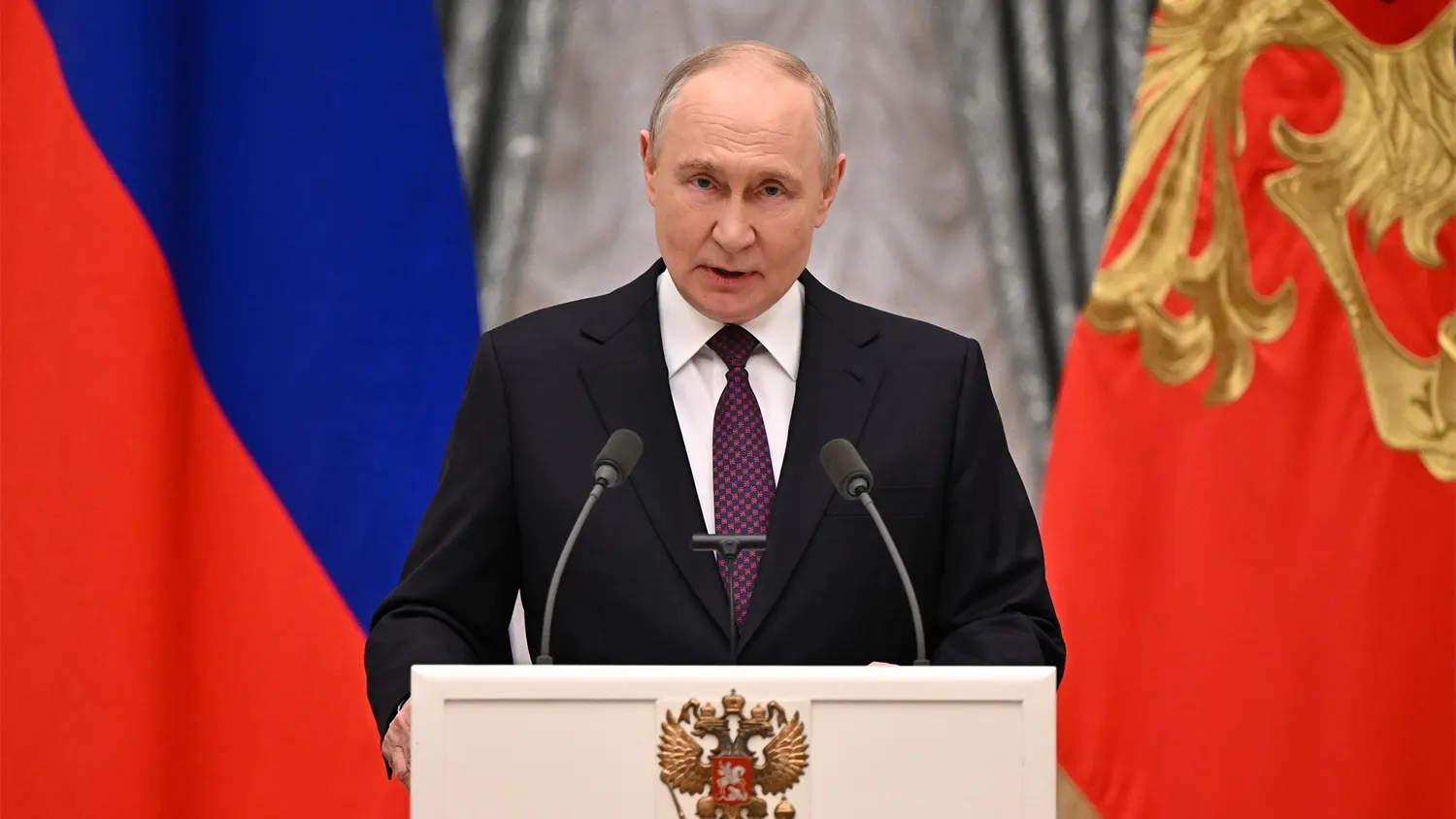 Russian President Vladimir Putin speaking at a podium with microphones, flanked by the Russian flag and state coat of arms.