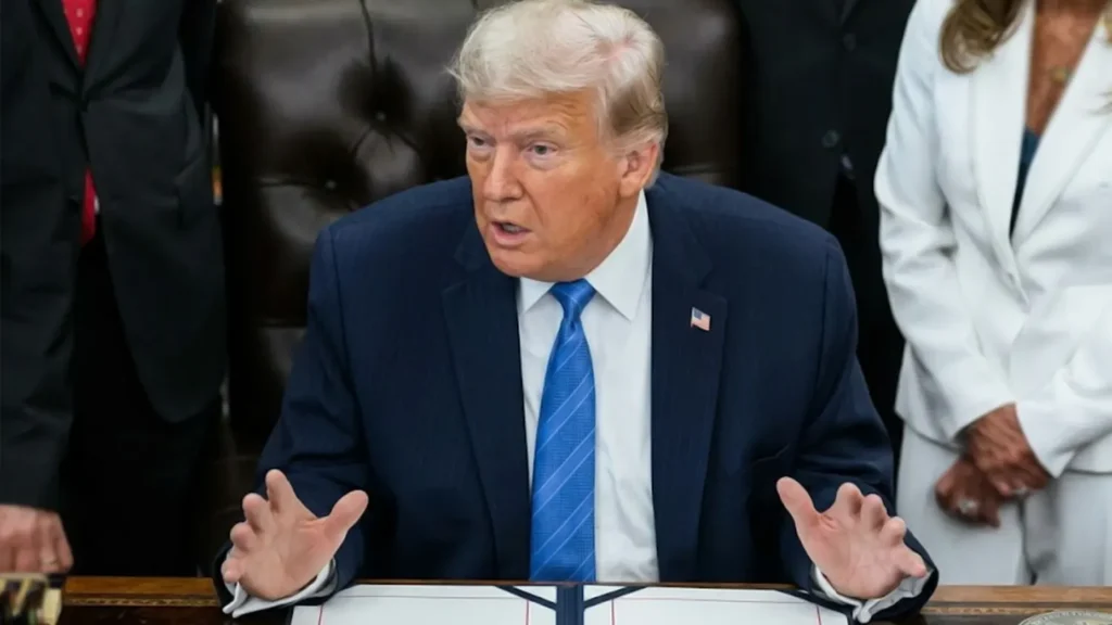 Close-up shot of Donald Trump seated at a wooden desk gesturing with both hands while discussing the USMCA trade agreement, flanked by officials.
