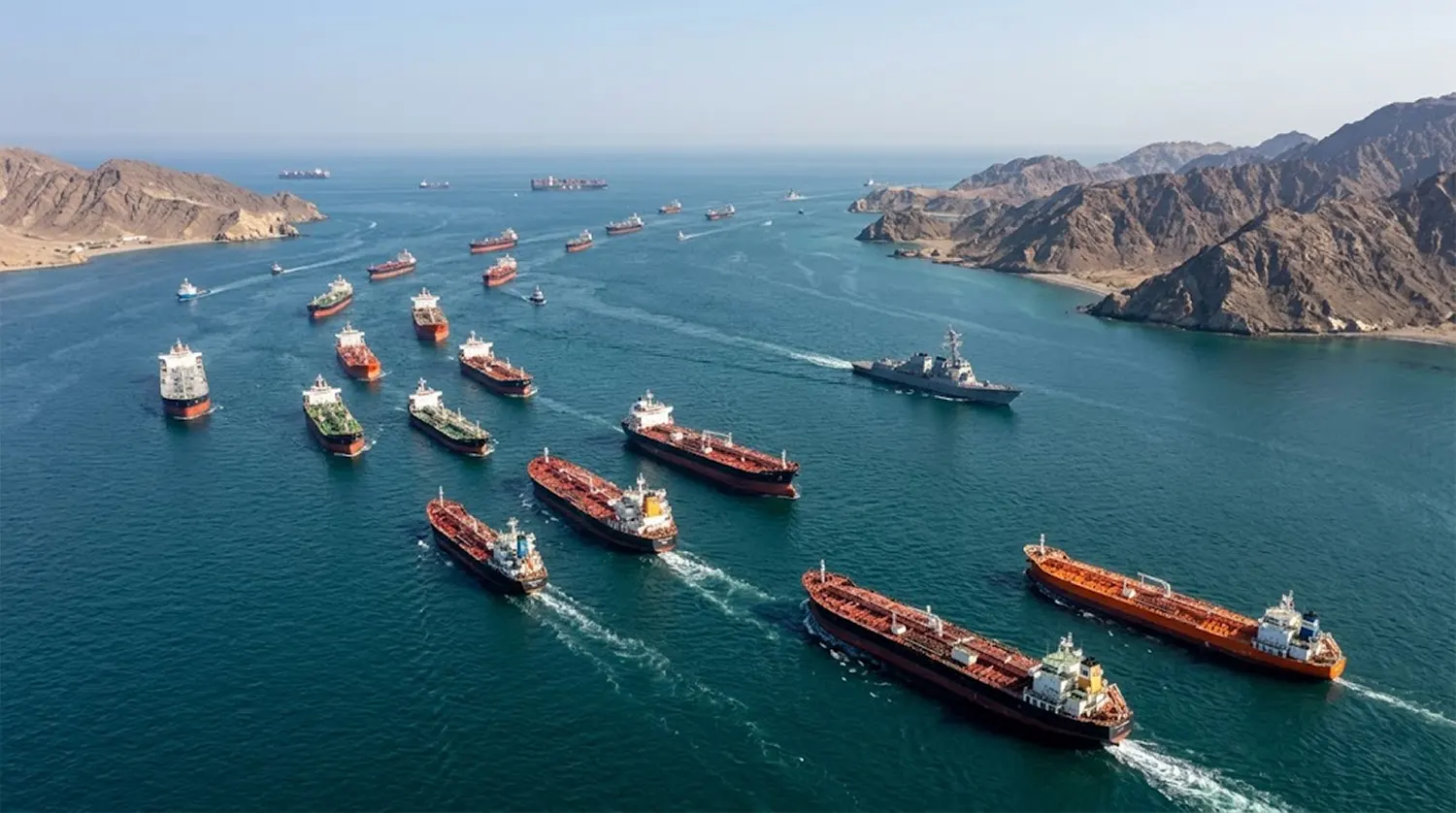 High-angle aerial view of commercial oil tankers and cargo ships moving through the Strait of Hormuz alongside a U.S. Navy destroyer following the 2026 U.S.-Iran peace framework agreement.