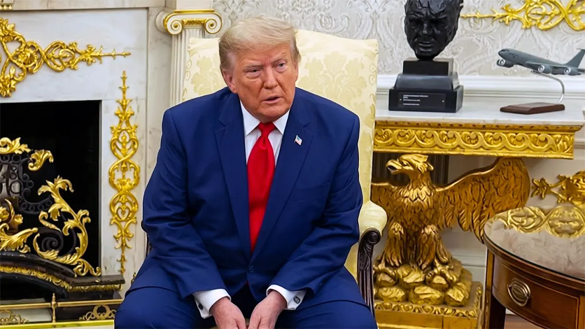 Donald Trump sitting in a chair inside the Oval Office wearing a blue suit and red tie, in a high-resolution portrait.