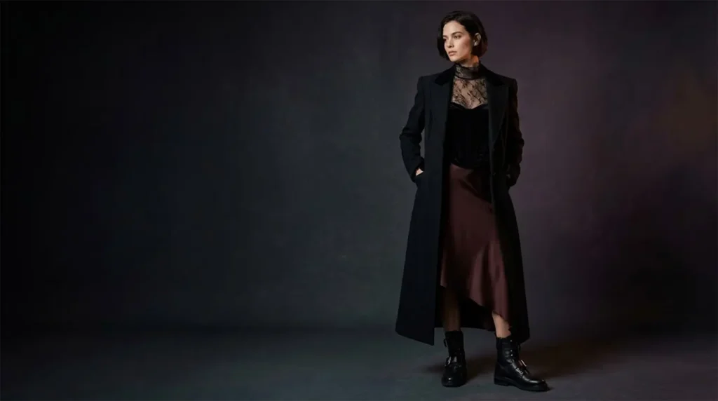 Model in a floor-length black wool duster coat, sheer lace high-neck blouse, and oxblood satin skirt in a moody studio.