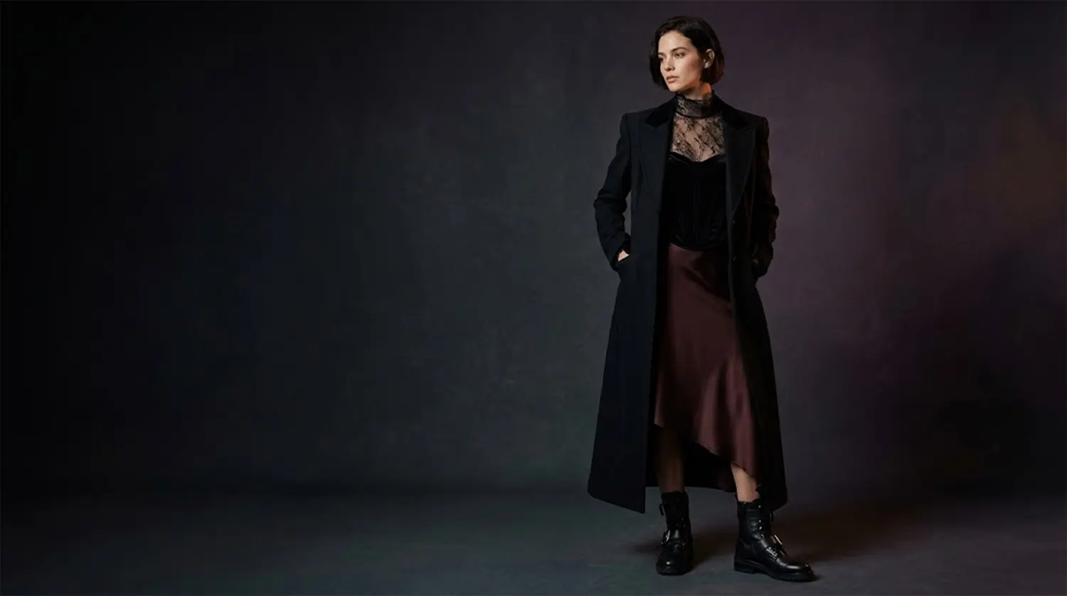 Model in a floor-length black wool duster coat, sheer lace high-neck blouse, and oxblood satin skirt in a moody studio.