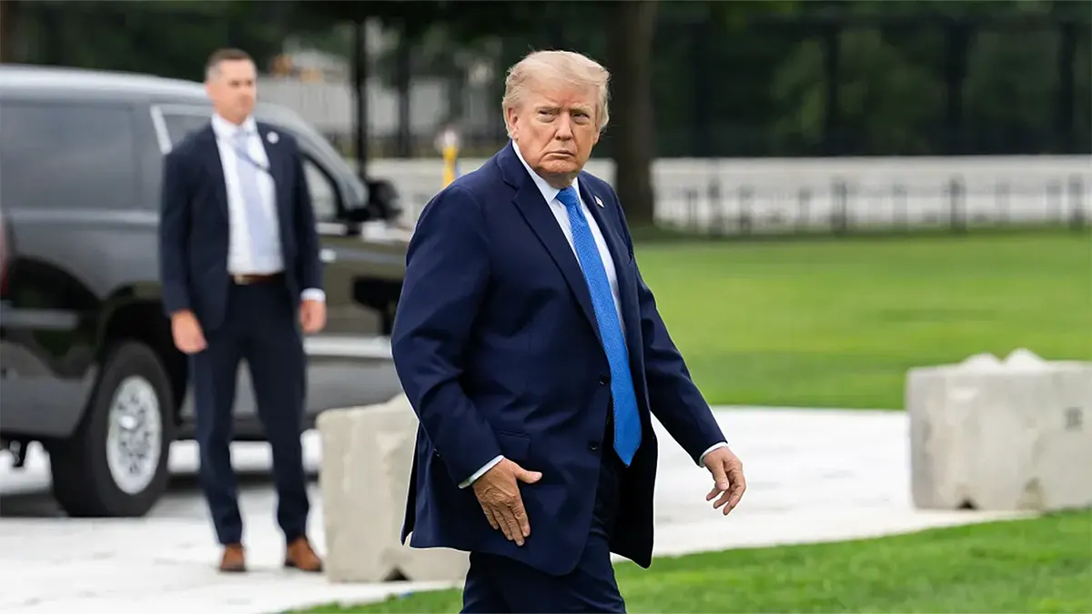Donald Trump wearing a dark suit and blue tie walking on the White House grounds near a black Secret Service SUV.