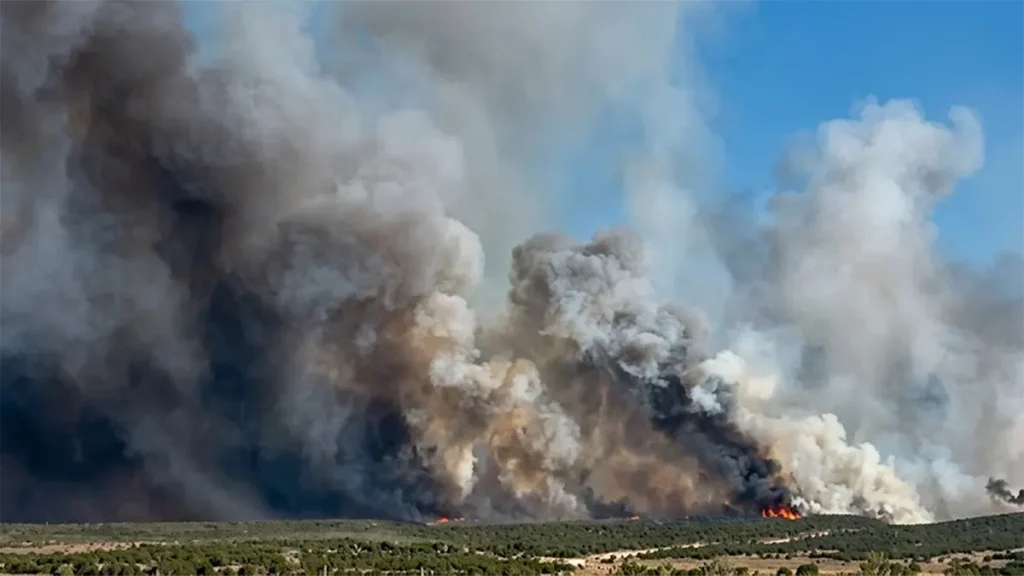 Ultra high-resolution wide shot of dense dark smoke billowing from a Utah wildfire spreading across a brush-covered landscape under a blue sky.